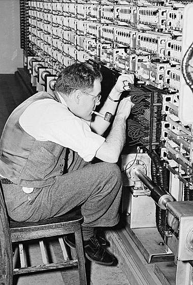 Black-and-white photograph of a technician in a waistcoat, pipe in mouth, working on the wiring of a tall relay-switching rack.
